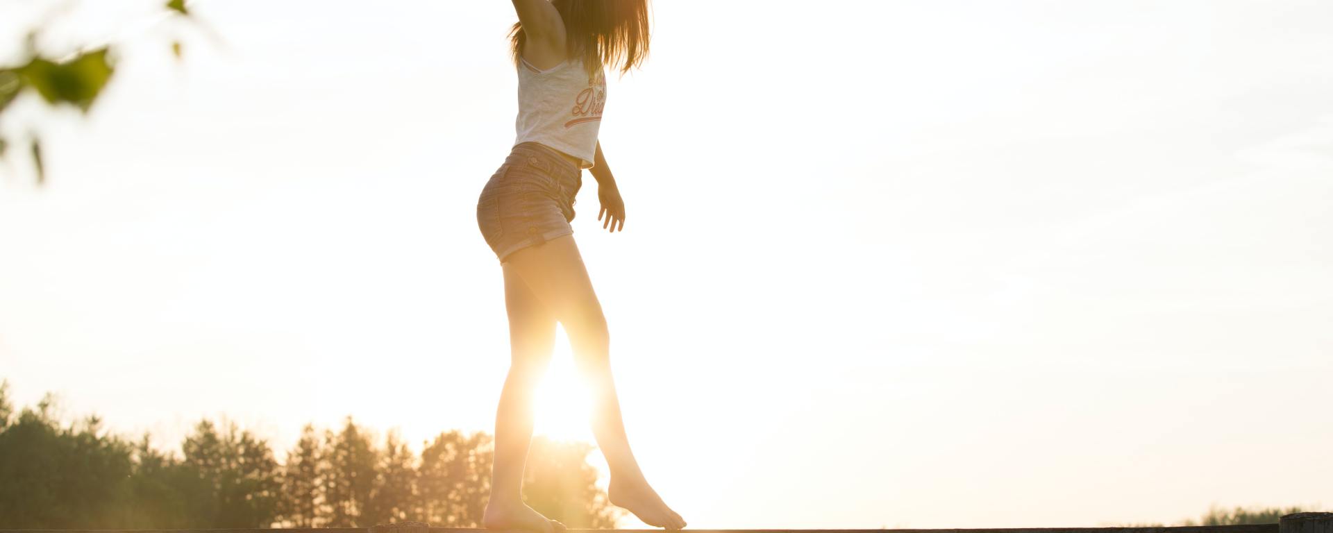 woman in a tank top and shorts walking across a wooden fence like a balance beam with the sun and trees in the background