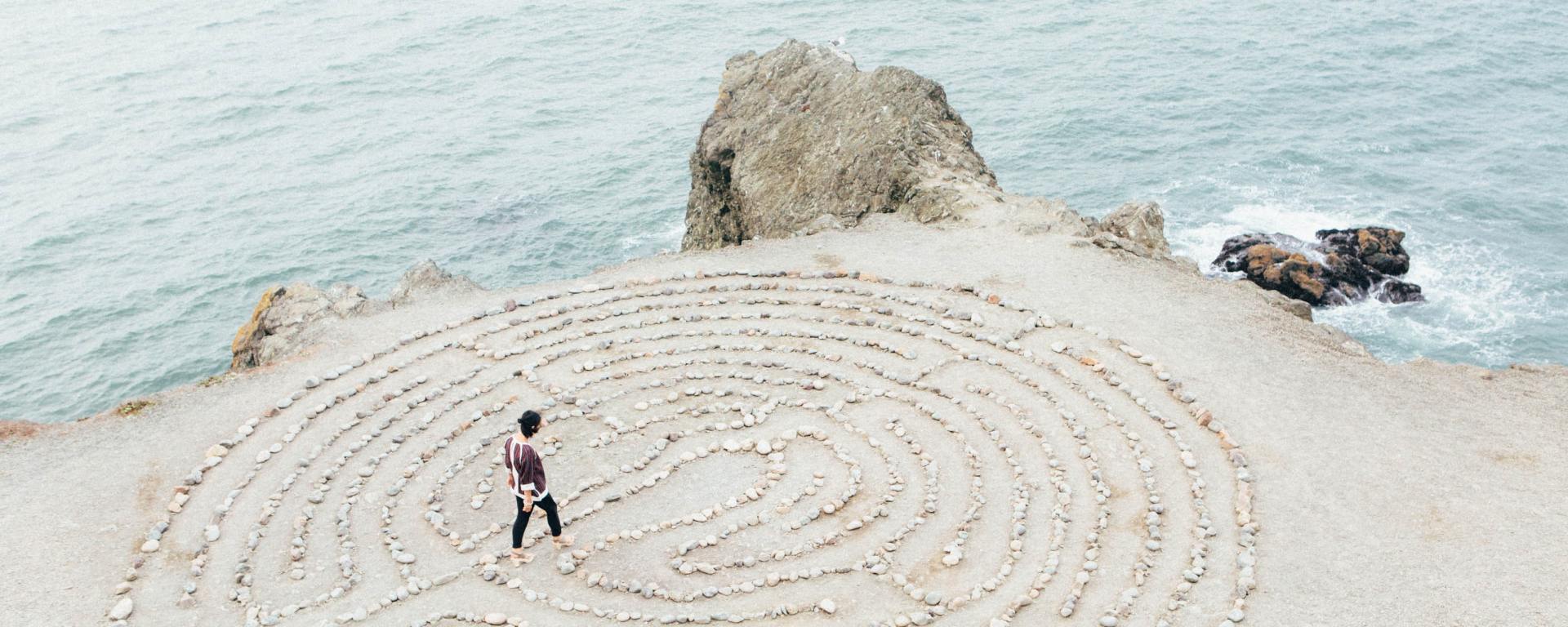 person walking through a circular stone maze in the sand by the ocean