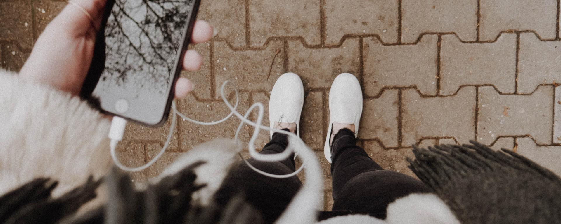 image facing down to someone's white shoes while they hold an iphone with headphones