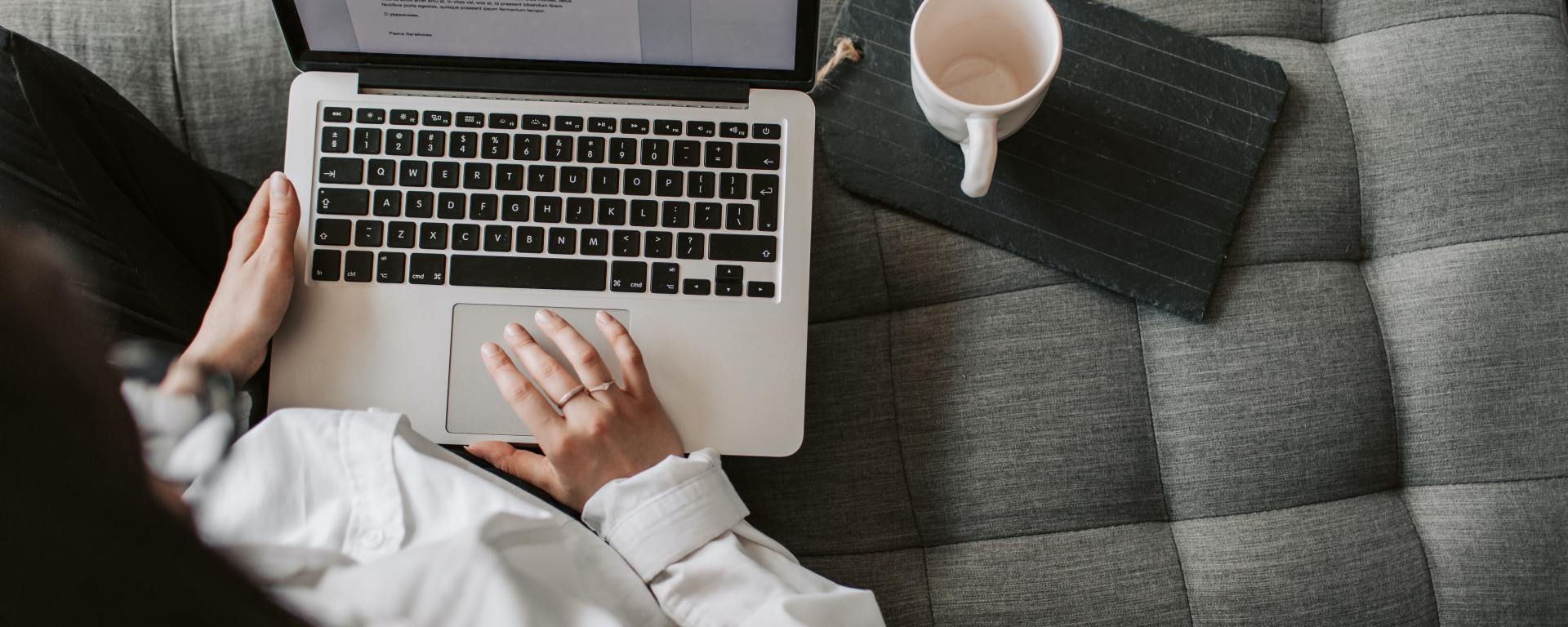 woman sitting on couch with computer in lap typing with coffee next to her
