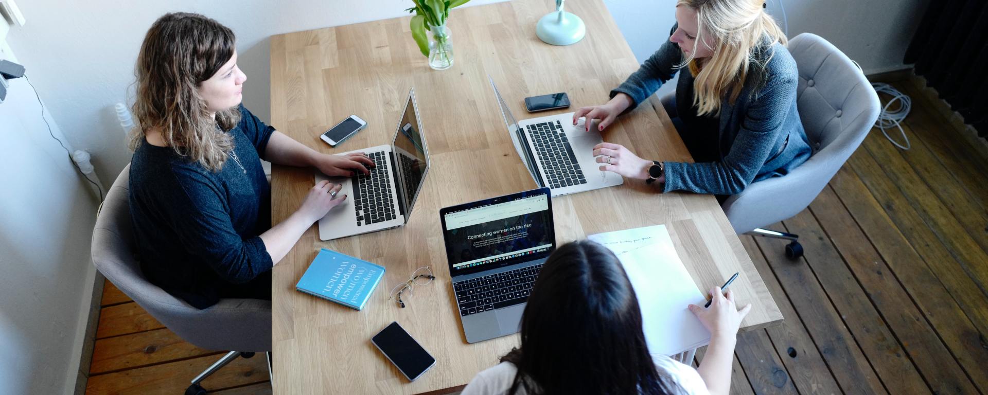 three women sitting at a table talking with laptops in front of them