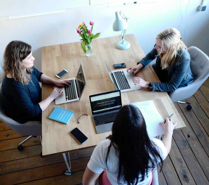 three women sitting at a table talking with laptops in front of them