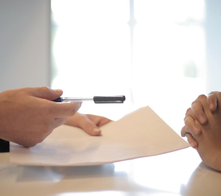 someone holding a piece of paper and a pen sitting across the table from someone with hands clasped on the table