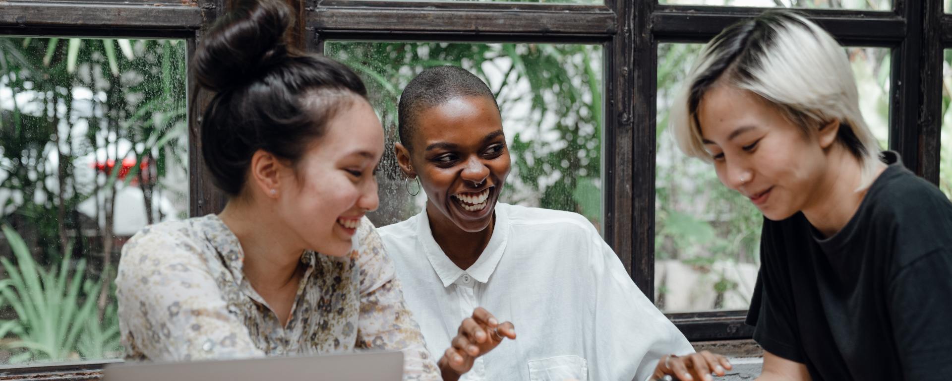 three young people at a table all smiling and laughing one with a laptop in front of them, one holding a phone